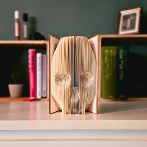 May include: A folded book sculpture in the shape of a skull. The pages are a light tan colour, with the skull's features clearly visible. The book is open, displaying the detailed folds. A shelf with books and ornaments is in the background.
