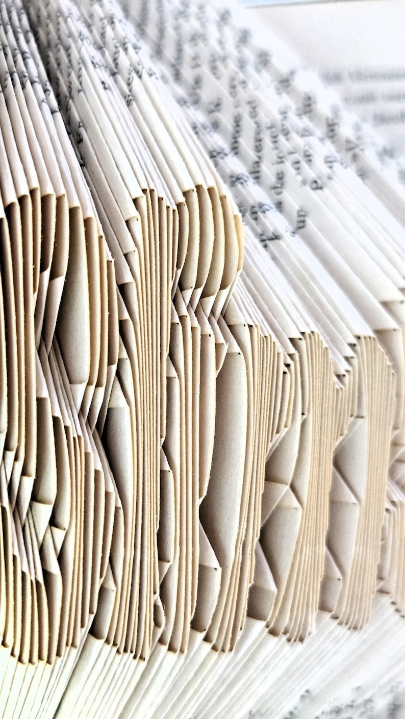 May include: A book with pages folded to create a three-dimensional sculptural effect. The off-white paper edges are precisely creased and layered to form structural patterns, revealing glimpses of printed black text on the individual page surfaces.
