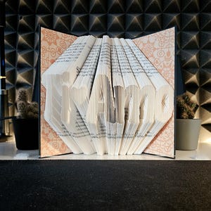 May include: A folded book art sculpture displaying the text Ti Amo, created by precisely folding the individual pages of a book. The interior covers feature a light peach-coloured pattern with intricate white floral and geometric line designs.