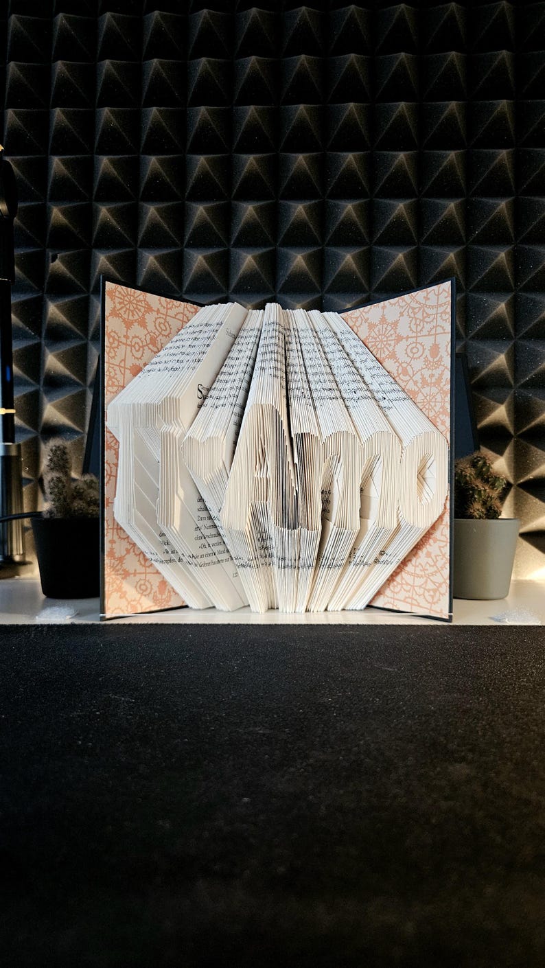 May include: A folded book art sculpture displaying the text Ti Amo, created by precisely folding the individual pages of a book. The interior covers feature a light peach-coloured pattern with intricate white floral and geometric line designs.