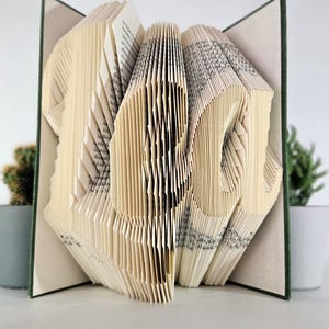 May include: A book with folded pages forming the word "LOL" in a three-dimensional sculpture. The book has a dark green cover, and the individual pages are creased and manipulated to create the letters, which are visible from the front view.
