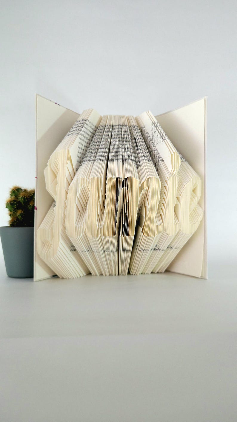 May include: A book sculpture with pages folded to form the name Jamie in a clear, legible font. The book is open, displaying the intricate paper folding technique used to create the three-dimensional lettering from the existing book pages.