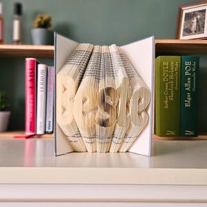 May include: A book sculpture spelling out "HOME", created by folding the pages. The open book is the focal point, with the letters standing out. Other books and a framed photo are visible in the background.