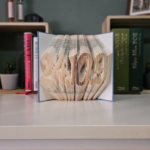 May include: A handcrafted book art sculpture featuring the numbers "2023" folded into the pages. The sculpture is displayed on a white surface, with books and decorative items in the background. The book has a blue border.