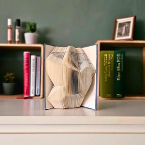 May include: A book sculpture with folded pages, forming a geometric shape. The open book sits on a white surface, with a green wall behind it. Wooden shelves display books and decorative items.