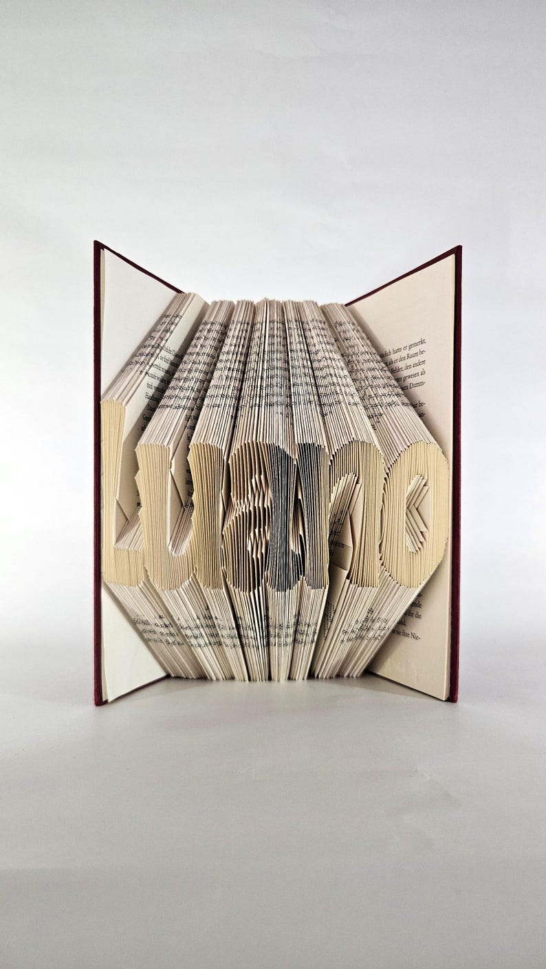 May include: A book with folded pages forming the name Luano. The book has a dark red cover and cream-coloured paper pages with black printed text. The pages are precisely creased and folded to create the three-dimensional letters of the name.