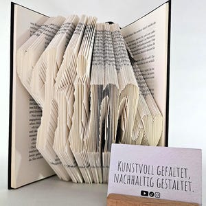 May include: A book with pages folded to form the word KIM, featuring black text on white paper. Beside the book is a small rectangular card in a wooden stand with the text KUNSTVOLL GEFALTET, NACHHALTIG GESTALTET and social media icons.