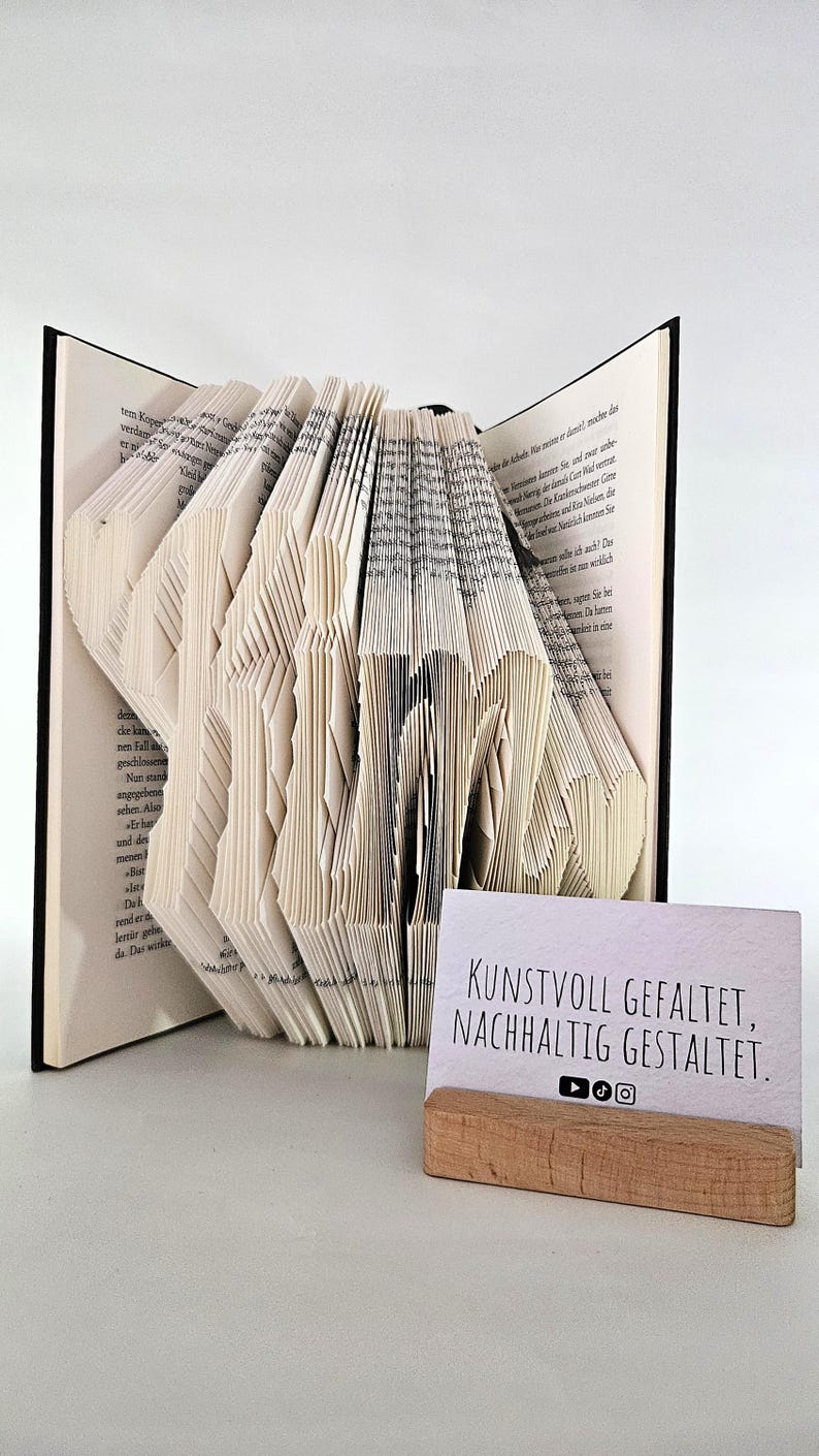 May include: A book with pages folded to form the word KIM, featuring black text on white paper. Beside the book is a small rectangular card in a wooden stand with the text KUNSTVOLL GEFALTET, NACHHALTIG GESTALTET and social media icons.
