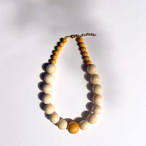 May include: A wooden bead necklace with a gold-colored chain and clasp. The necklace features a mix of light beige and yellow-orange beads, with a larger orange bead in the center. The beads are round and smooth.