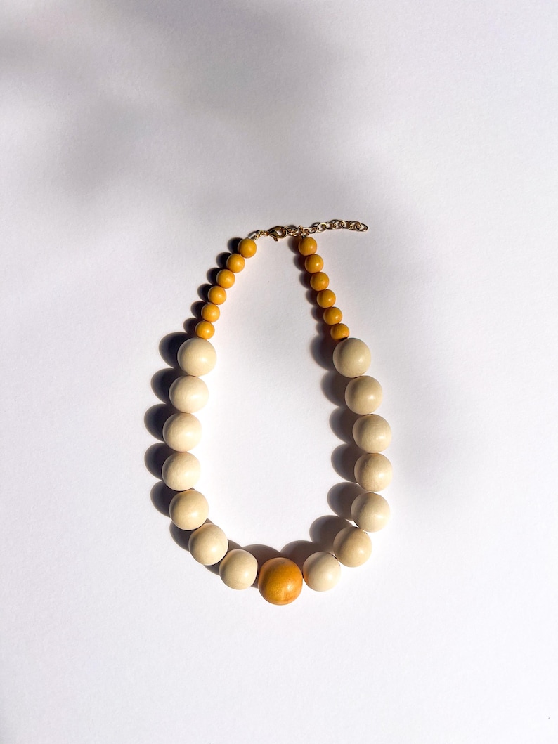 May include: A wooden bead necklace with a gold-colored chain and clasp. The necklace features a mix of light beige and yellow-orange beads, with a larger orange bead in the center. The beads are round and smooth.