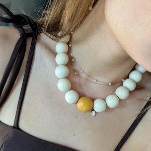 May include: A cream-colored beaded necklace with a large yellow bead and a delicate gold chain necklace with small pearls. The necklace is worn over a dark brown top.