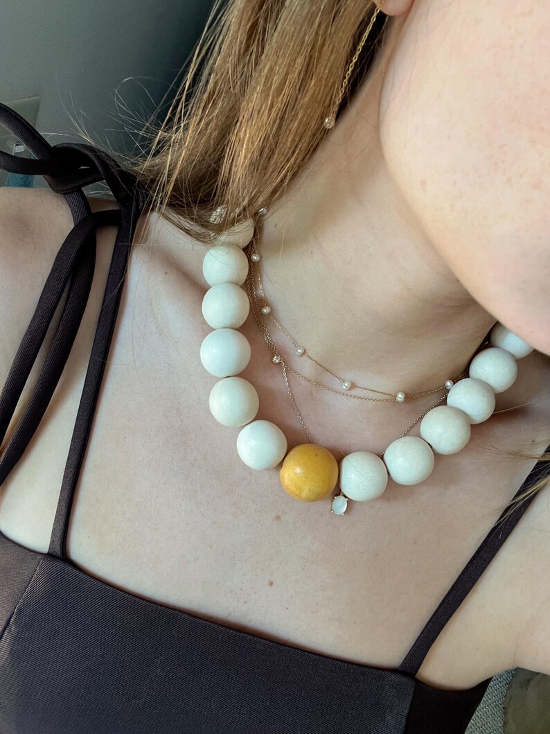 May include: A cream-colored beaded necklace with a large yellow bead and a delicate gold chain necklace with small pearls. The necklace is worn over a dark brown top.
