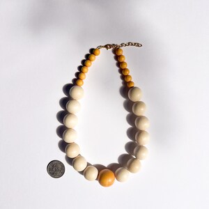 May include: A wooden bead necklace featuring large, cream-colored beads and smaller, light brown beads. The necklace has a gold-colored chain and clasp. A single orange bead is centered in the design. The necklace is shown on a white background.