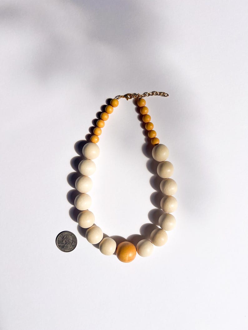 May include: A wooden bead necklace featuring large, cream-colored beads and smaller, light brown beads. The necklace has a gold-colored chain and clasp. A single orange bead is centered in the design. The necklace is shown on a white background.