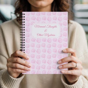 May include: A spiral-bound notebook with a light pink cover featuring a seashell pattern. The notebook has the text "Mermaid Thoughts & Other Mysteries" in a decorative font within a white frame. The notebook is held open.