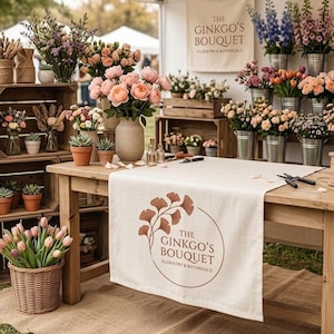 May include: A floral market stall featuring a wooden table with a runner displaying the text The Ginkgo's Bouquet Floristry & Botanicals. The setup includes various potted plants, bouquets of pink roses, tulips, and dried flowers in wooden crates.