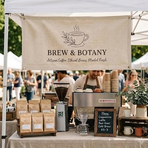 May include: A beige fabric banner with the text BREW & BOTANY, Artisan Coffee. Ethical Beans. Market Fresh. and a coffee cup graphic. Below, a coffee grinder, bags of coffee beans, a glass dripper, and a menu board on a table.