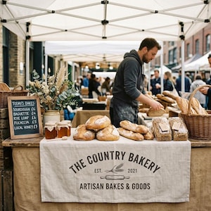 May include: A bakery stall display featuring a beige linen tablecloth with the text THE COUNTRY BAKERY EST. 2021 ARTISAN BAKES & GOODS, loaves of bread, jars of honey, a chalkboard menu, and a wicker basket under a white canopy tent.