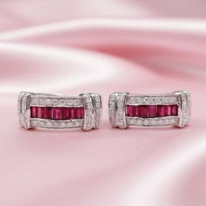 May include: Two silver earrings with a rectangular design. Each earring has a central row of rectangular, deep red gemstones, framed by smaller, clear gemstones. The earrings are displayed on a soft pink background.