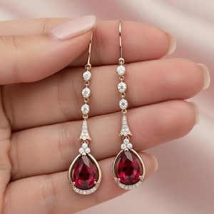 May include: Gold-toned drop earrings featuring teardrop-shaped, deep red gemstones. The earrings have a delicate design with small, clear accent stones and a long, slender chain. The gemstones are surrounded by a halo of smaller stones.