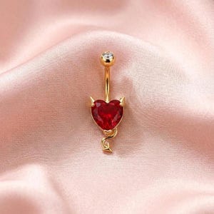 May include: Gold-colored belly button ring with a red heart-shaped charm. The charm has small horns and a tail. The top of the ring has a small, clear gem. The background is a pink satin fabric.