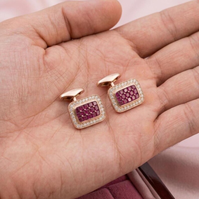 May include: A pair of rectangular rose gold cufflinks featuring a central grid of small square-cut red rubies surrounded by a border of tiny clear white diamonds. The cufflinks have a polished metal finish and a classic hinged toggle backing.