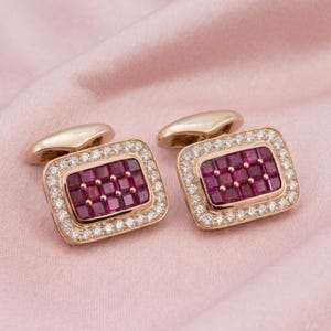 May include: A pair of rectangular cufflinks crafted from rose gold, each featuring a central grid of deep red square-cut rubies surrounded by a border of small, clear, round-cut diamonds, set in a polished metal frame with a hinged toggle back.