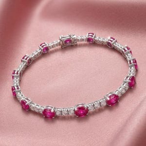 May include: A silver-toned tennis bracelet featuring a repeating pattern of oval-cut vibrant red gemstones alternating with small, clear, round-cut stones. The bracelet includes a secure metal clasp mechanism and a polished, reflective finish.