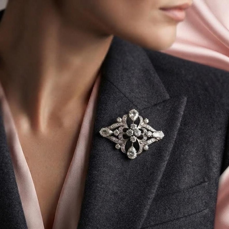 May include: A silver-toned brooch featuring an intricate openwork design set with multiple clear, faceted stones. The piece has a symmetrical, elongated diamond shape with pear-shaped stones at the points and round stones throughout the center.