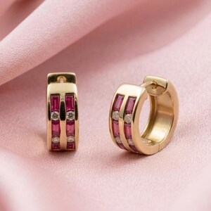 May include: A pair of gold hoop earrings featuring two parallel rows of rectangular baguette-cut red gemstones alternating with small round clear stones. The earrings have a hinged closure mechanism and a polished metallic finish throughout.