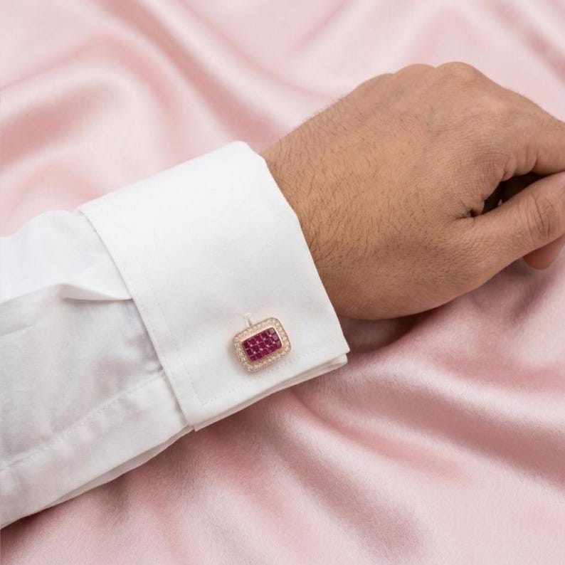 May include: A rectangular cufflink featuring a grid of small, deep red square-cut stones set in a metallic frame, surrounded by a border of tiny clear stones, attached to the white fabric cuff of a long-sleeved shirt.