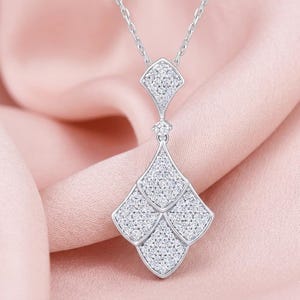 May include: A silver pendant necklace with a geometric design. The pendant features a series of diamond-like shapes, each filled with small, sparkling stones. The necklace hangs on a delicate silver chain, set against a pink background.