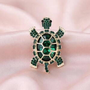 May include: A gold-toned turtle brooch featuring a large, faceted dark green central stone on the shell, surrounded by smaller, sparkling green rhinestones covering the head, legs, and the outer border of the shell, creating a detailed animal design.