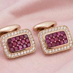 May include: A pair of rectangular cufflinks crafted from rose gold metal. Each cufflink features a central grid of deep red square-cut rubies surrounded by a border of small, clear, round-cut white stones set in a polished metal frame.
