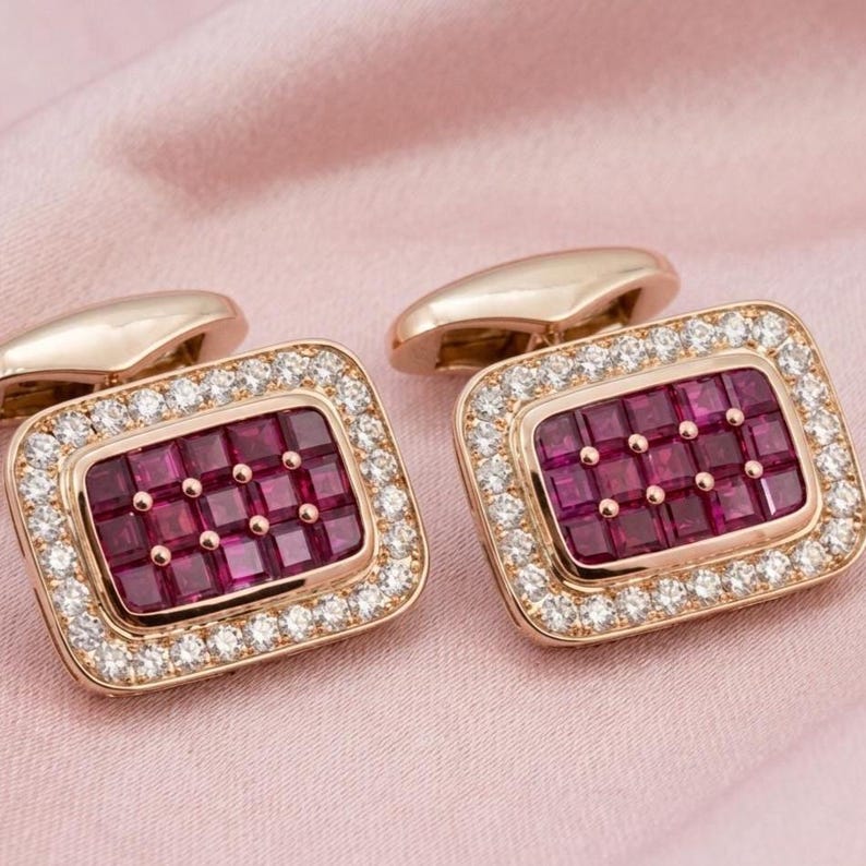 May include: A pair of rectangular cufflinks crafted from rose gold metal. Each cufflink features a central grid of deep red square-cut rubies surrounded by a border of small, clear, round-cut white stones set in a polished metal frame.