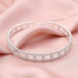 May include: A silver-toned choker necklace featuring two parallel rows of clear, sparkling rhinestones connected by small, square-shaped rhinestone links, creating an open, geometric pattern throughout the entire length of the jewelry piece.