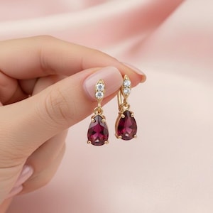 May include: A pair of gold-colored drop earrings with a teardrop-shaped, deep red gemstone and small, clear accent stones. The earrings are held up against a soft pink background.
