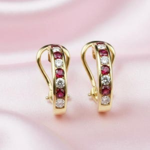 May include: A pair of gold hoop earrings with a row of alternating red and white gemstones. The earrings are set against a soft pink background, highlighting the jewelry's sparkle and design.