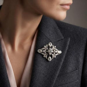 May include: A silver-toned brooch featuring an intricate openwork design adorned with multiple clear, faceted stones. The piece includes a central cluster of round stones surrounded by pear-shaped and smaller round stones in a symmetrical pattern.