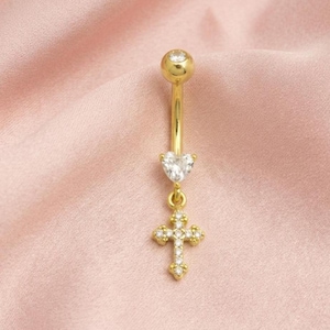 May include: Gold-colored belly button ring with a clear crystal heart and cross pendant. The top ball and cross are adorned with small, clear crystals. The jewelry is set against a pink background.
