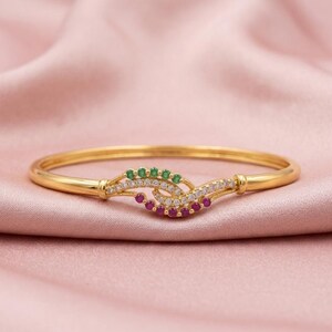 May include: A gold bangle bracelet featuring a central wave-shaped design. The ornament is embellished with a row of small green stones, a row of clear white stones, and a row of small pink stones arranged in a curved, layered pattern.