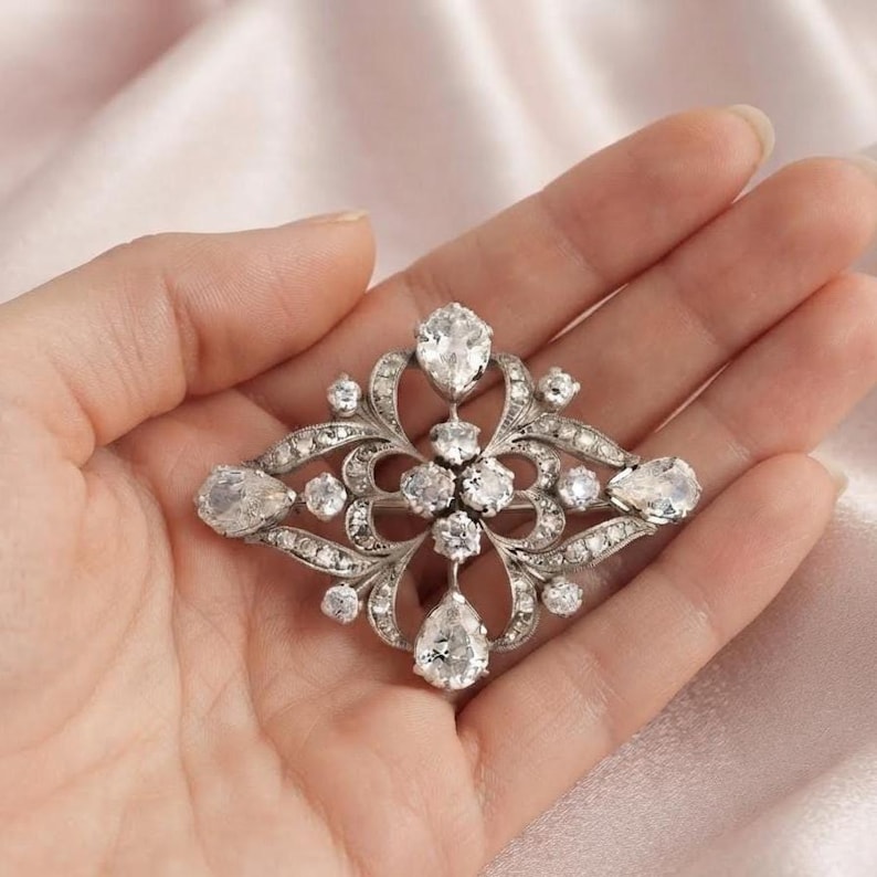 May include: A silver-toned brooch featuring a symmetrical, openwork metal design adorned with multiple clear, faceted stones. The arrangement includes four prominent pear-shaped stones at the cardinal points and smaller round stones throughout.