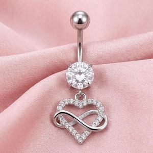 May include: A silver belly button ring with a large, clear, round gemstone at the top and a dangling heart-shaped charm. The charm features a heart design intertwined with an infinity symbol, both encrusted with small, clear gemstones.