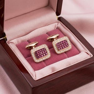 May include: A pair of rectangular cufflinks featuring a grid of deep red square stones set in gold-toned metal, surrounded by a border of small clear stones. The cufflinks are displayed inside a dark wooden box with a soft pink velvet interior.