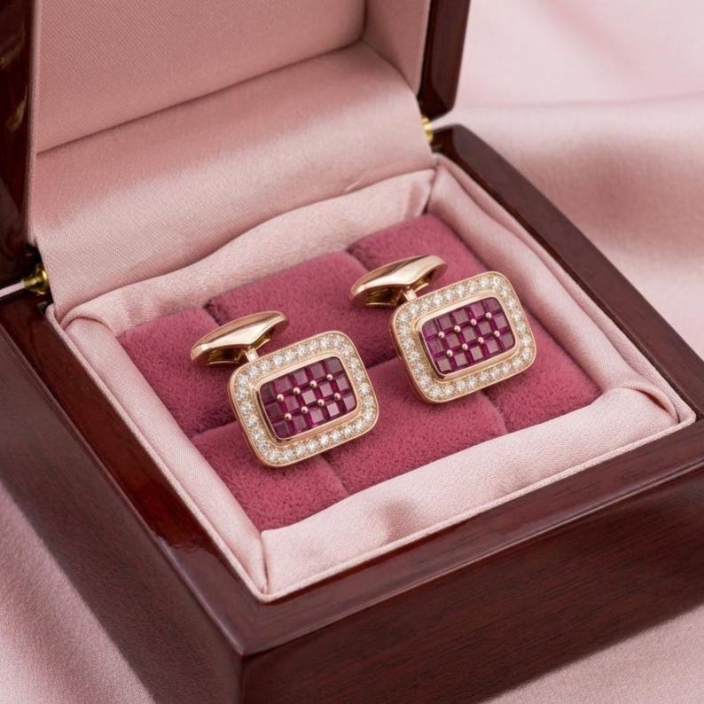 May include: A pair of rectangular cufflinks featuring a grid of deep red square stones set in gold-toned metal, surrounded by a border of small clear stones. The cufflinks are displayed inside a dark wooden box with a soft pink velvet interior.