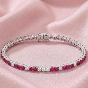 May include: A silver-toned tennis bracelet featuring a continuous row of alternating round-cut red rubies and clear white diamonds. The piece is secured with a rectangular metal box clasp mechanism that connects the two ends of the circular band.
