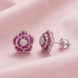 May include: A pair of stud earrings shaped like five-petaled flowers. Each earring features a central cluster of small white stones surrounded by concentric layers of vibrant pink stones set in a polished silver-toned metal with a post backing.