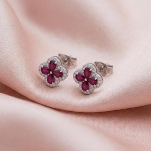 May include: A pair of silver stud earrings featuring a four-leaf clover design. Each earring contains four pear-cut deep pink stones arranged in a floral pattern, surrounded by a border of small, clear, round-cut white stones.