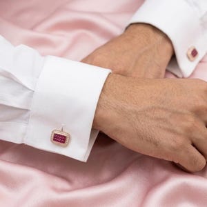 May include: A pair of rectangular cufflinks featuring a grid of small, deep red stones set within a metallic frame, secured on the white fabric cuffs of a long-sleeved shirt. The cufflinks have a polished finish and a border of tiny clear stones.