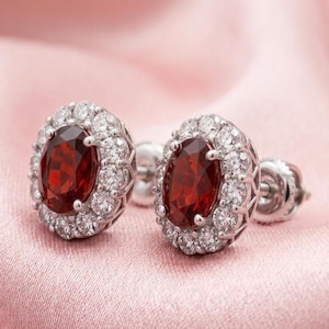 May include: A pair of stud earrings featuring oval-cut deep red garnet gemstones at the center. Each garnet is surrounded by a halo of small, round, clear white diamonds set in polished silver-toned metal with secure post backings.